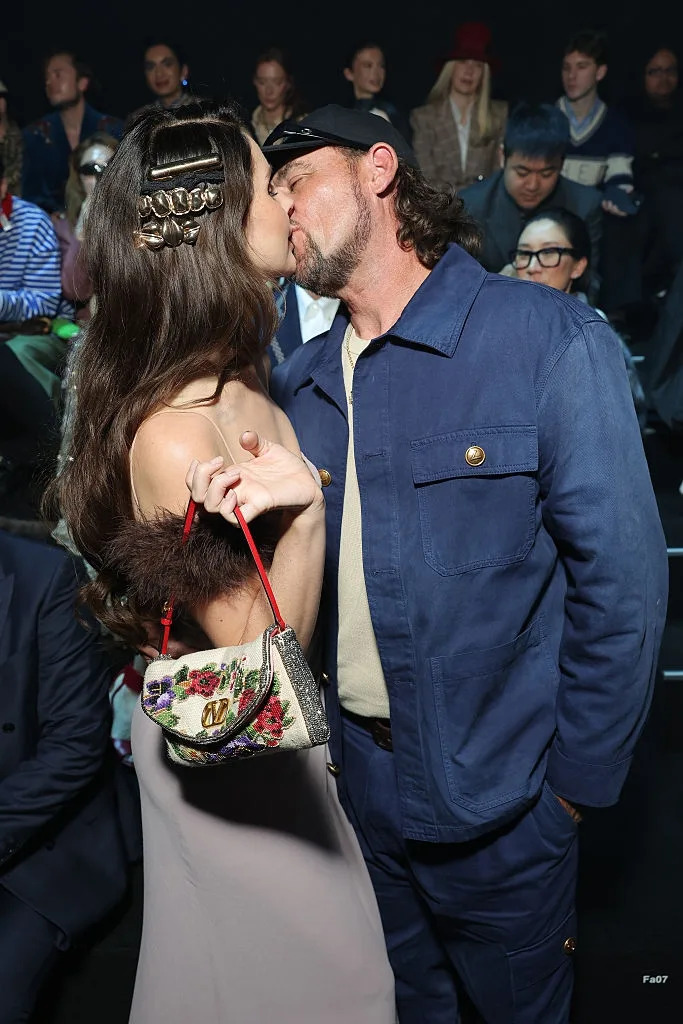 A couple, one in a glamorous outfit with hair clips and a handbag, share a kiss at a fashion event, surrounded by an audience