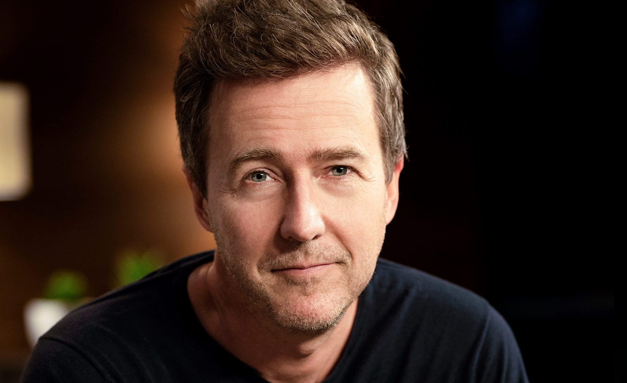 A man with short brown hair and light stubble, wearing a black shirt, looks directly at the camera with a slight smile. The background is softly blurred in warm tones.
