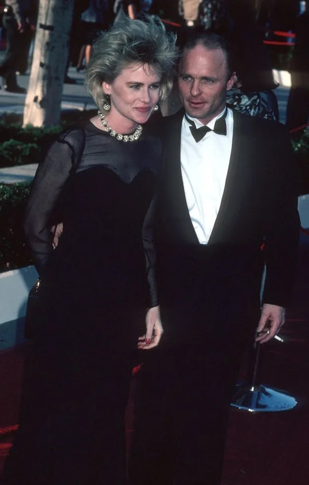 A man and woman pose on a red carpet. He wears a tuxedo; she wears an elegant black gown with a statement necklace