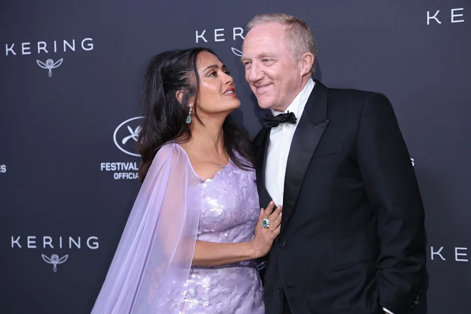 A couple on a red carpet event; the woman in an elegant gown with sheer shawl and the man in a classic tuxedo, smiling and embracing