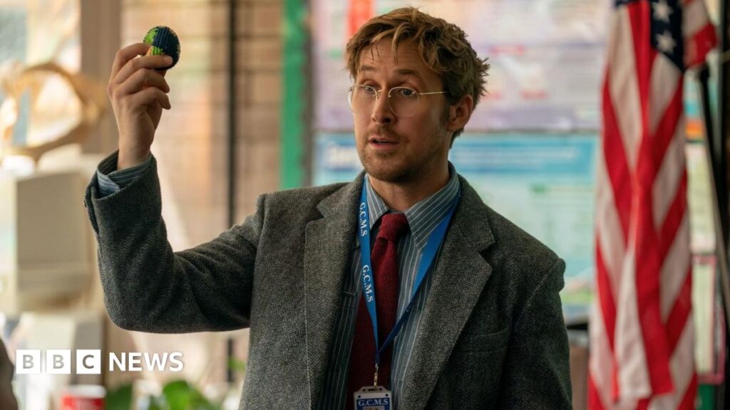 Picture of Ryan Gosling in a still from Project Hail Mary. He is wearing a suit jacket, shirt, tie and glasses and is holding a small globe toy.