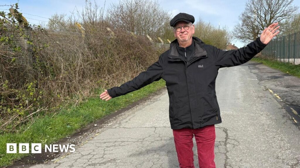 A middle-aged man wearing red trousers, a black jacket and a cap stands on an empty lane smiling with his arms outstretched.