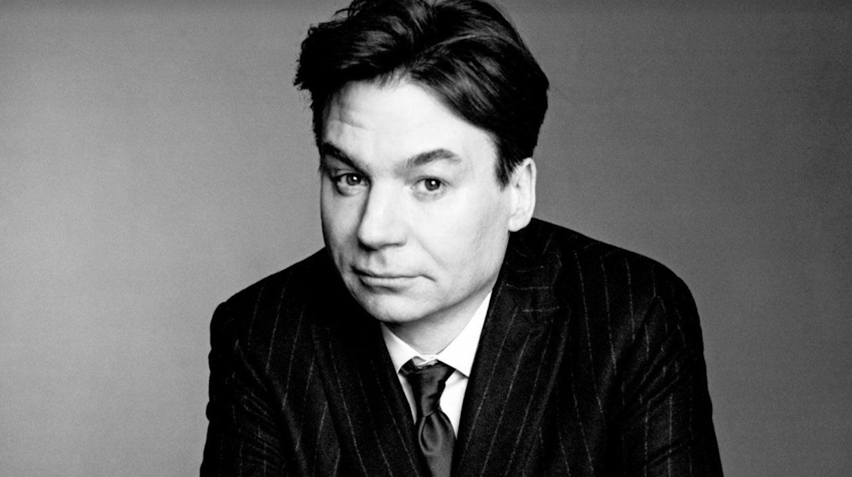 A man with dark hair in a pinstripe suit and tie looks directly at the camera with a slight, knowing smile against a plain background. The image is in black and white.