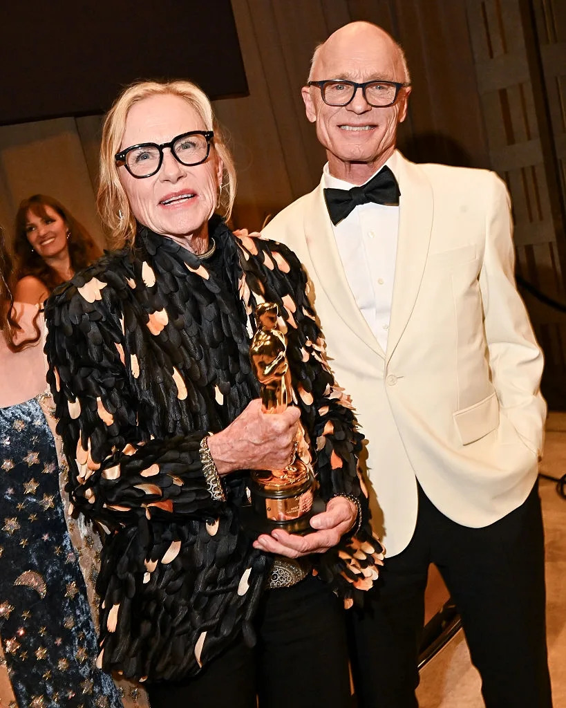 Two individuals pose at a formal event. One holds a statue. The other is in a tuxedo. Both wear glasses