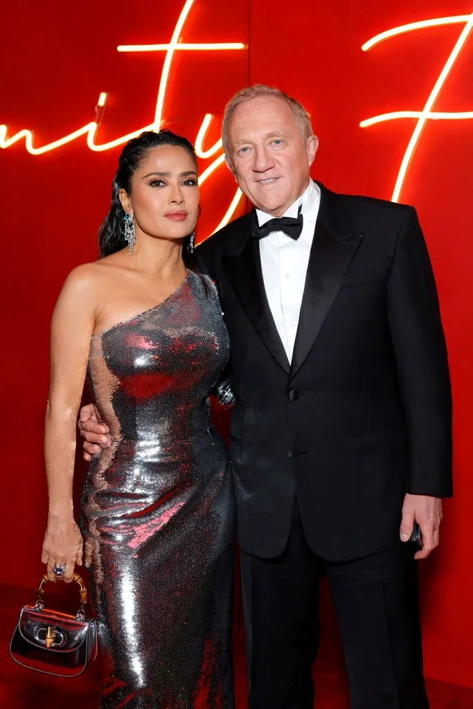 I don't know who these individuals are, but a woman in a metallic one-shoulder gown and a man in a tuxedo pose together at a formal event
