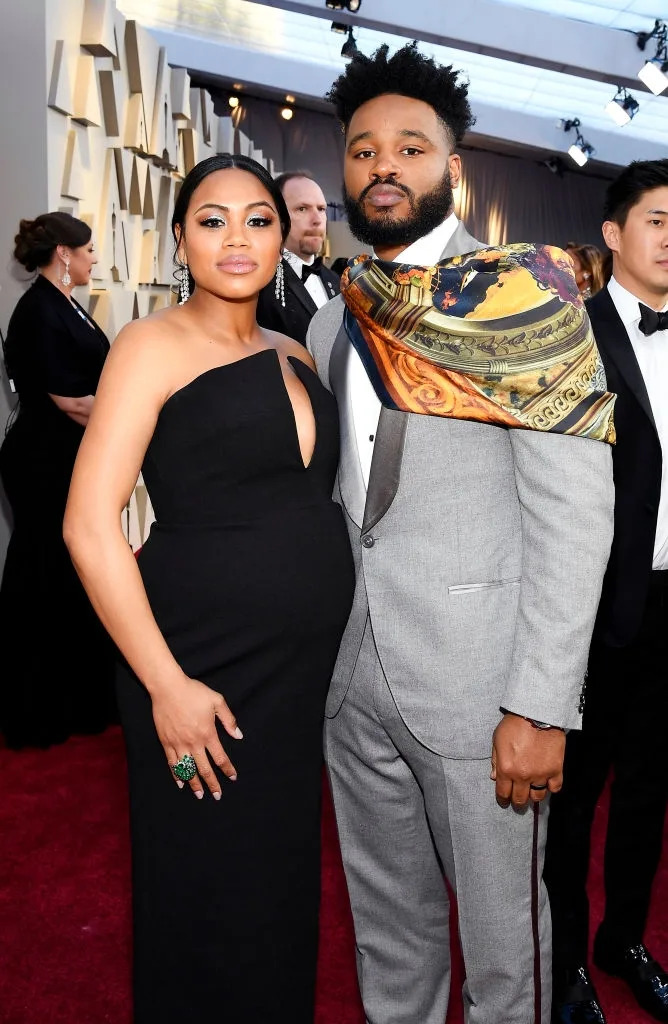 A couple poses on a red carpet. The woman wears an elegant black strapless gown, while the man dons a gray suit with a patterned scarf