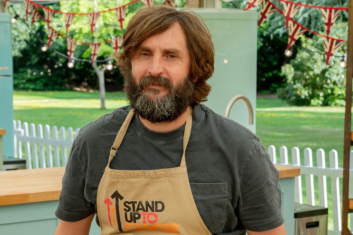 Joe Wilkinson looking ahead in the Bake Off kitchen, wearing a grey t-shirt and a light brown apron.