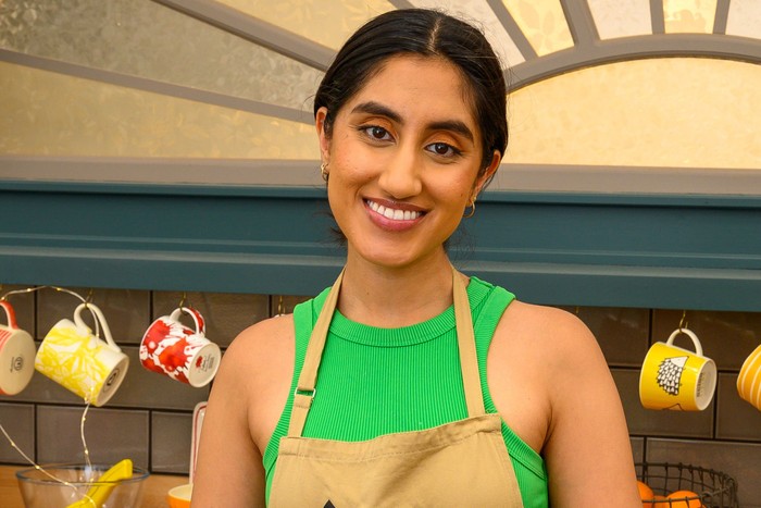 Ambika Mod smiling ahead, wearing a bright green top and a light brown apron.