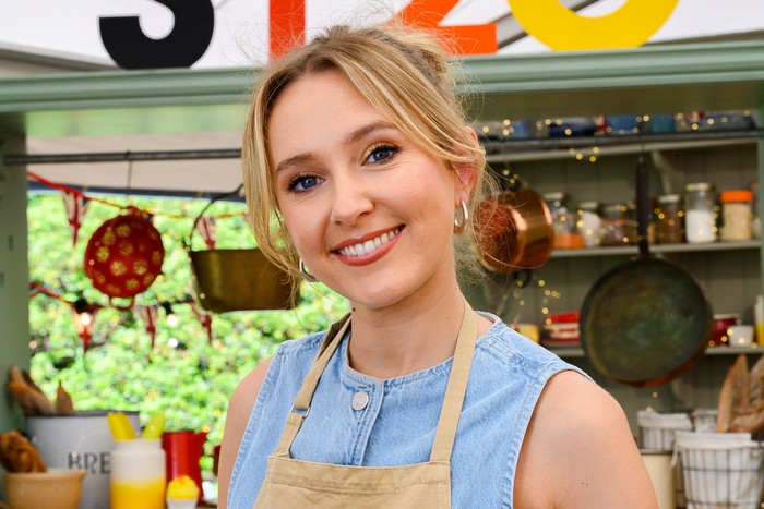 Rose Ayling-Ellis smiling ahead, wearing a light blue shirt and brown apron.