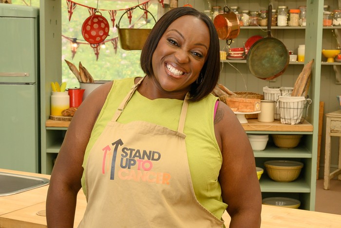 Judi Love smiling with her head bent to one side, wearing a green shirt and brown apron.