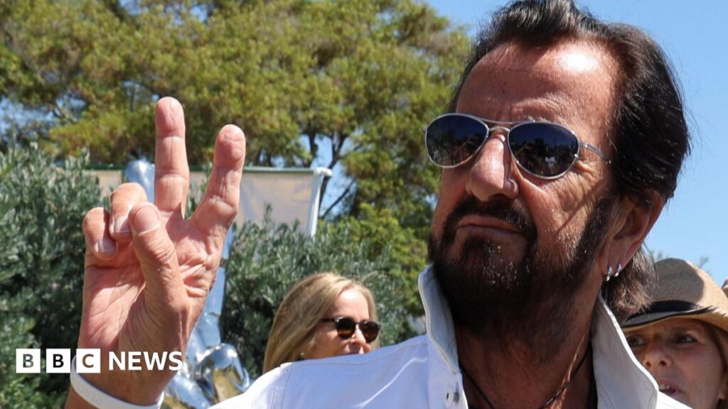 Ringo Starr, in his early 80s with black hair brushed back, a black beard and sunglasses. He is wearing a white linen shirt with the collar turned up and is holding up two fingers on his right hand in a peace symbol.