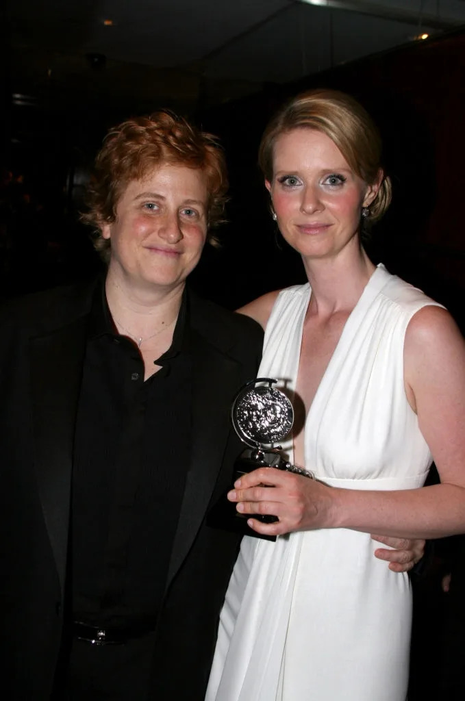 Two individuals pose together, one holding an award. The person on the right wears an elegant white dress