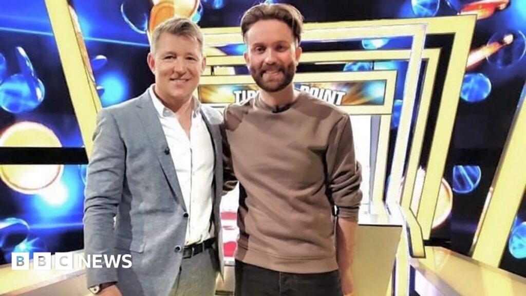 Publicity image of Ben Shephard and Jamie posing together on the Tipping Poin set, which has a blue background and a row of yellow square arches above.