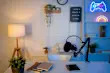 Modern cozy home office setup with microphone, notebooks, and plants on desk. Warm lamp lighting with colorful neon wall signs, creating stylish workspace for gaming, podcasting, or content creation.