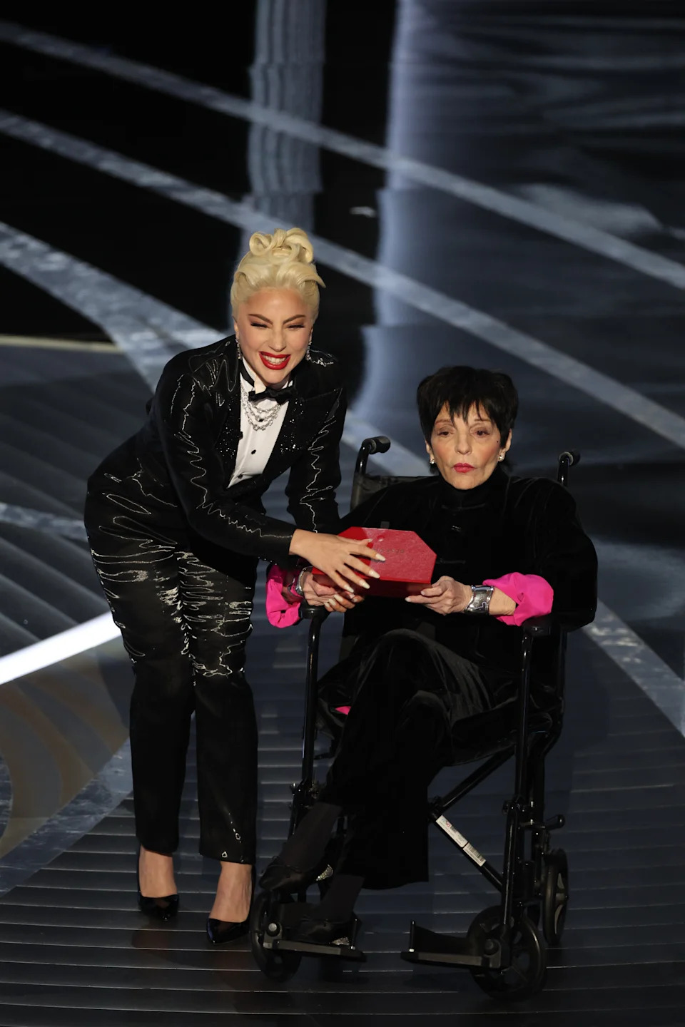 Two individuals on stage, one helping the other in a wheelchair. The standing person wears a shiny suit; the seated person wears a velvet outfit