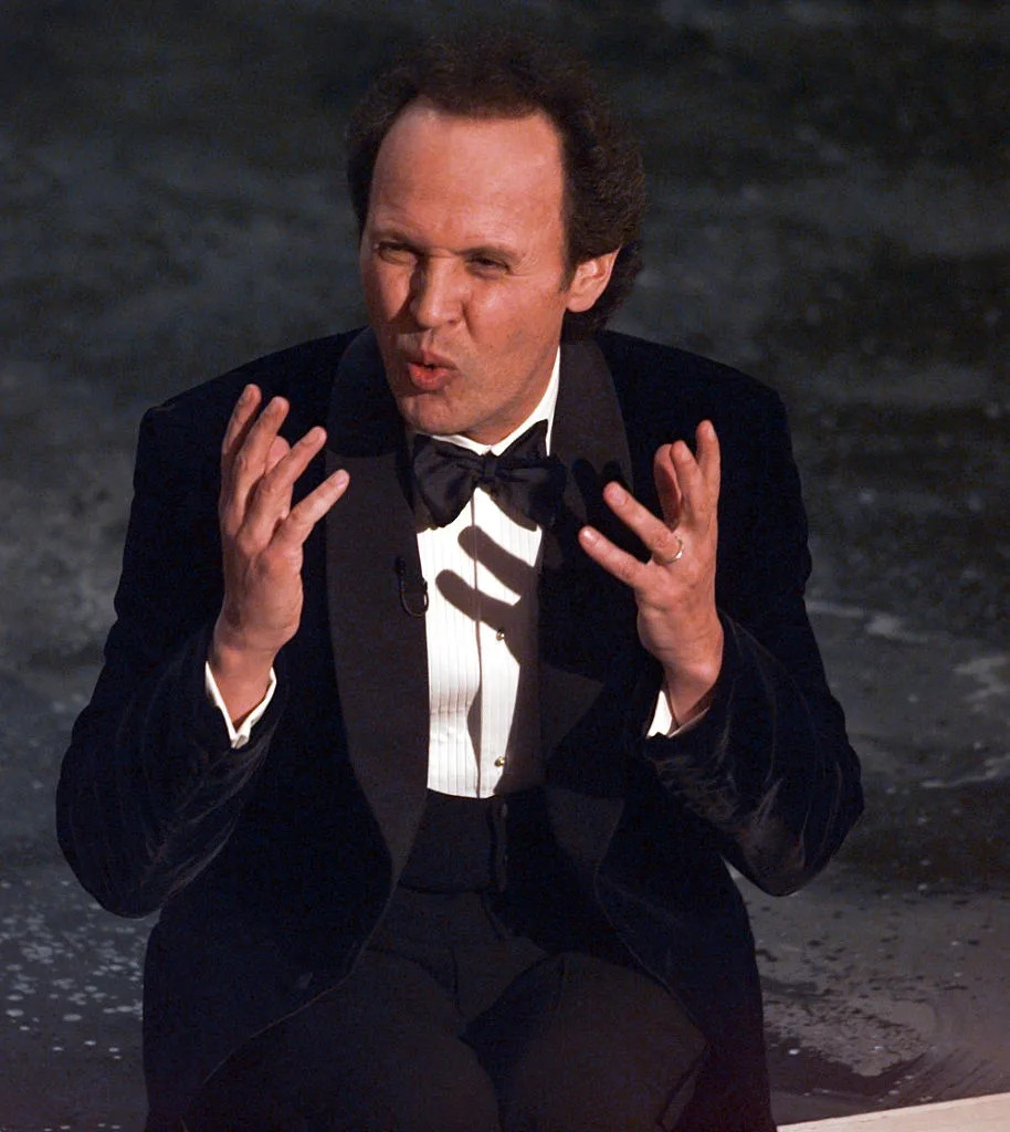 A person in a tuxedo gestures expressively on a stage, appearing to perform or speak animatedly
