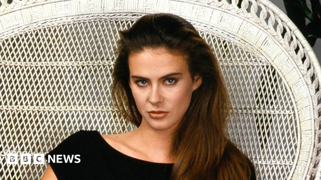 Annabel Schofield, a woman with long brown hair sits with in a white wicker chair, wearing a black dress. She looks directly at the camera with a serious expression.