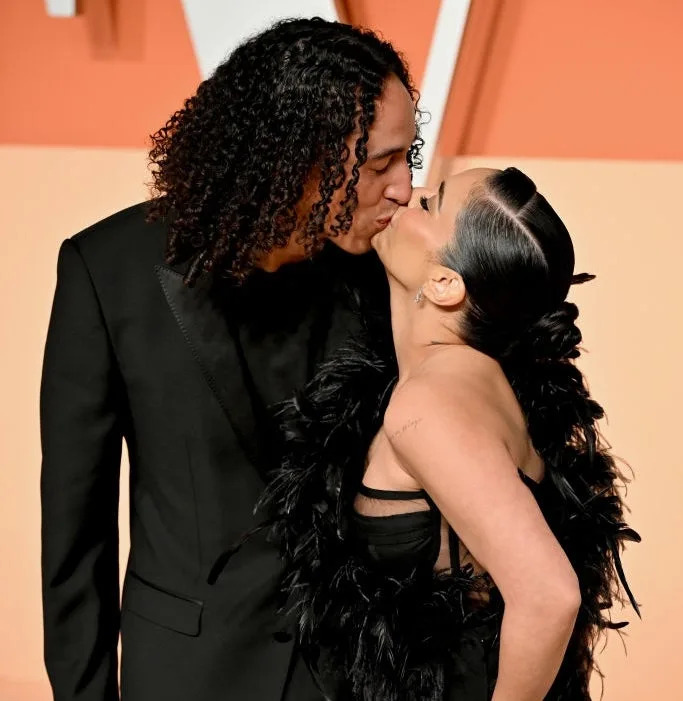 A couple kisses on the red carpet. One wears a black suit; the other wears a black dress with feather details and a thigh-high slit