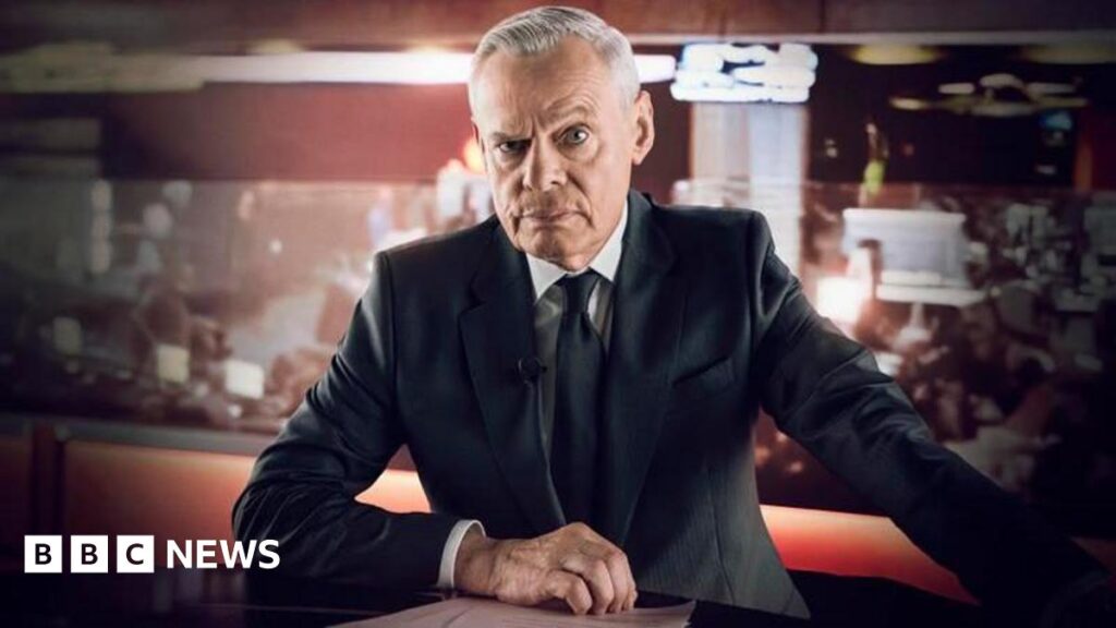 Clunes as Edwards. He wears a white shirt, with black suit jacket and tie. his right hand is rested on the desk in front of him, with his left reaching out