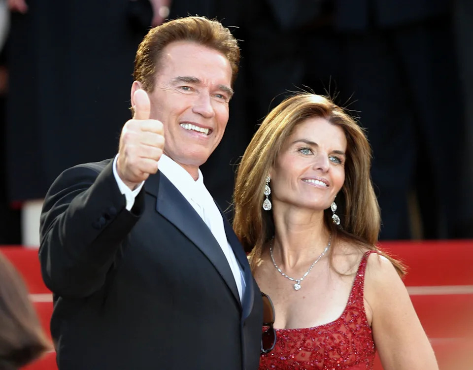 Two people on a red carpet, one in a suit giving a thumbs up, the other in an elegant sleeveless dress with sparkling earrings