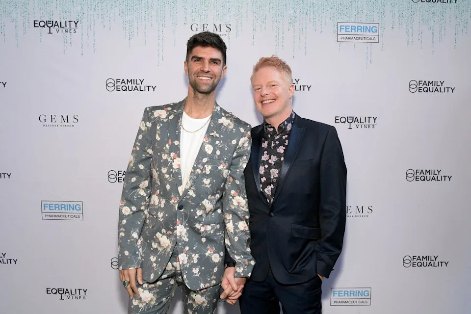 Two people on a red carpet, both smiling and holding hands. One wears a floral suit, the other a dark suit with a floral shirt. Background features sponsor logos