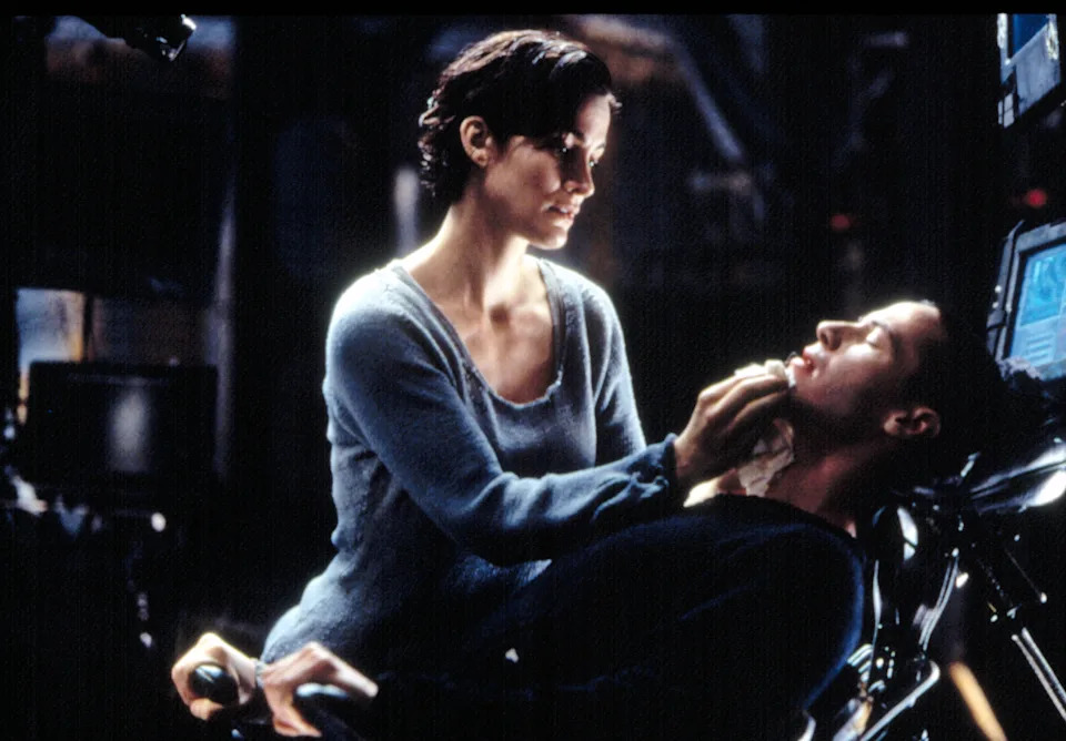 A woman gently touches a man's face as he reclines in a futuristic chair with screens, creating a tense, intimate moment in a sci-fi setting