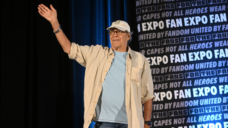 Chevy Chase waving and wearing a ball cap, blue shirt, and tan jacket