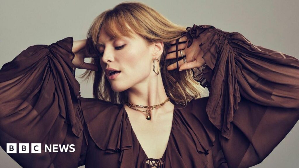 Freya Ridings holds her hands to her head in a promotional photo for her new music. She is dressed in a billowing, chocolate brown top with elaborate frills.