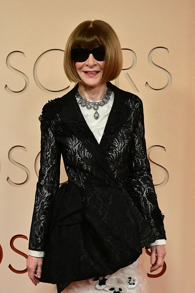 Person on red carpet in stylish black patterned jacket, statement necklace, and sunglasses