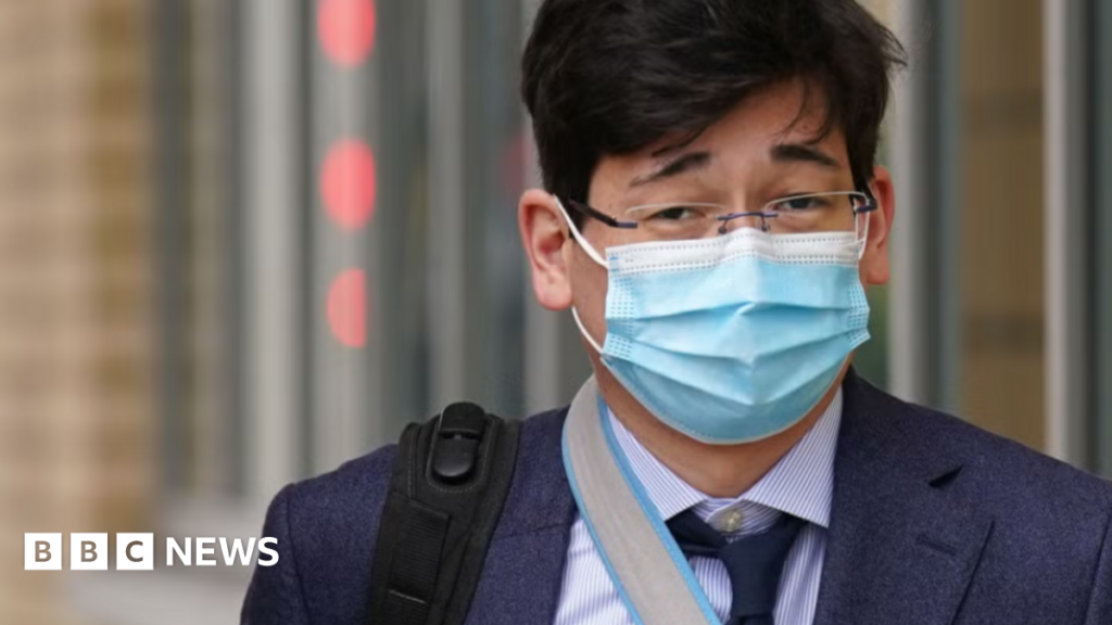 Jonathon Dean wearing a navy suit with a navy tie and a white shirt and glasses. He is walking outside of a court building with the strap of a black rucksack over one shoulder and a facemask on.