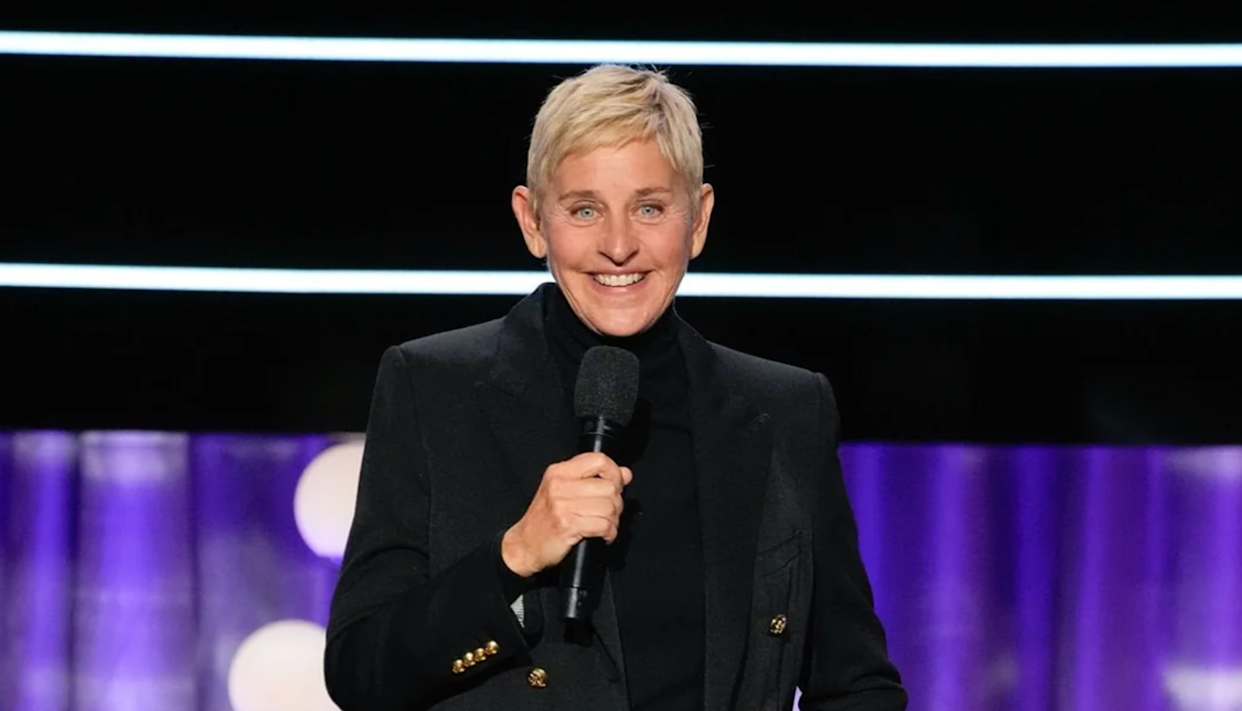 A person with short blonde hair wearing a black jacket and turtleneck holds a microphone and smiles on stage with purple lighting and horizontal light strips in the background.