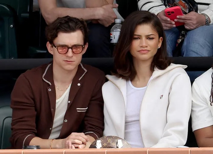 Three people seated at an event, with a woman wearing a white jacket in the center, flanked by men in casual attire