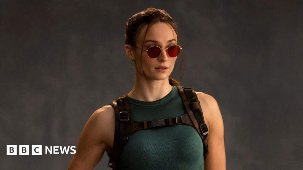A photo shows Sophie Turner as Lara Croft. She wears red-tinted sunglasses, her hair tied back. She wears a green vest top and tight, thigh-length shorts. One of her fingerless-gloved hands rests on a pistol in a thigh holster.