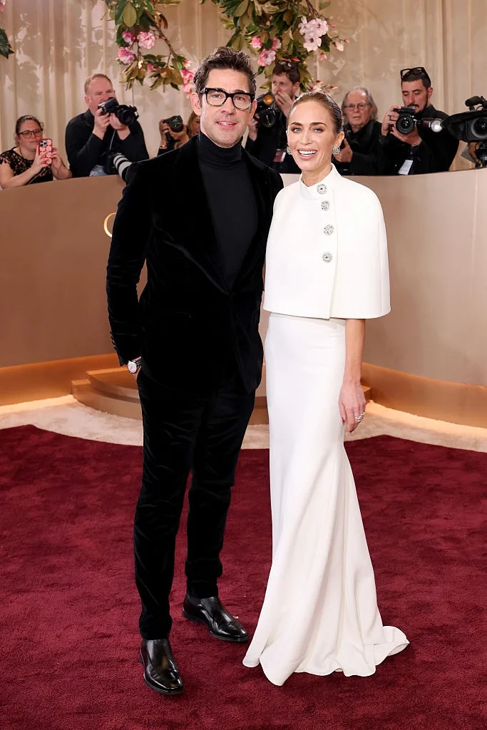 A couple poses on a red carpet; the man wears a black velvet suit, and the woman wears an elegant white gown with a cape and button details