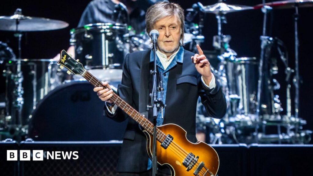 Paul McCartney on stage wearing a blue jacket, blue shirt and with a guitar. He is pointing one finger in the hair.