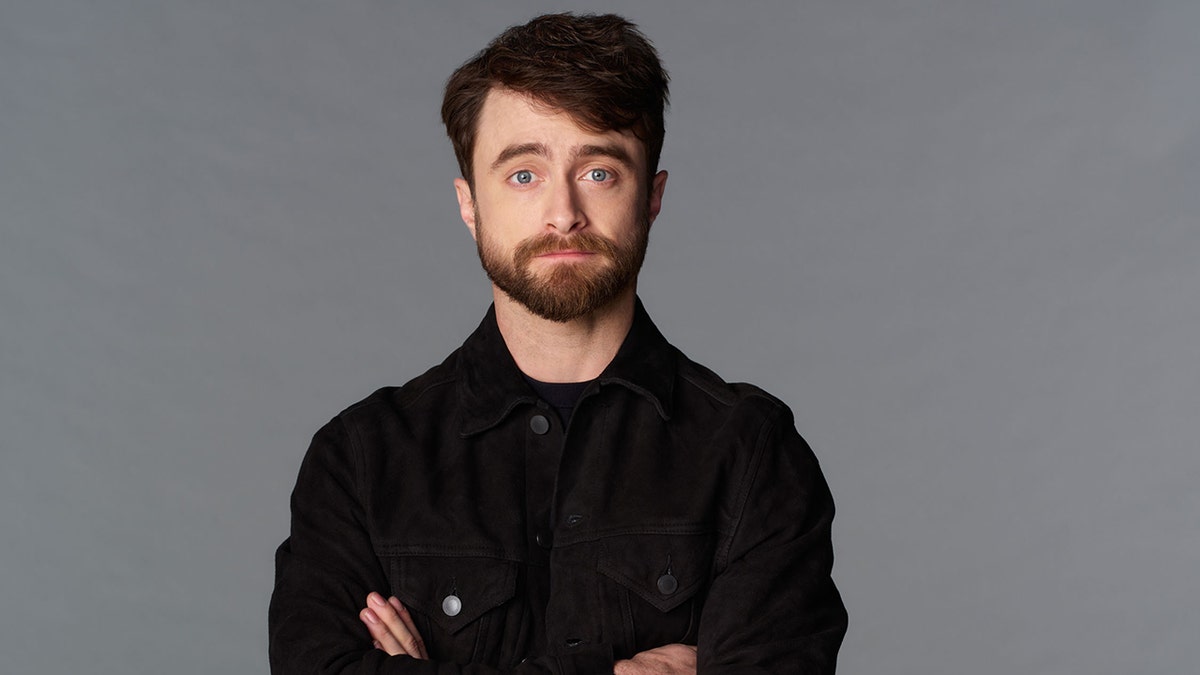 Daniel Radcliffe in a black shirt standing in front of a black background.