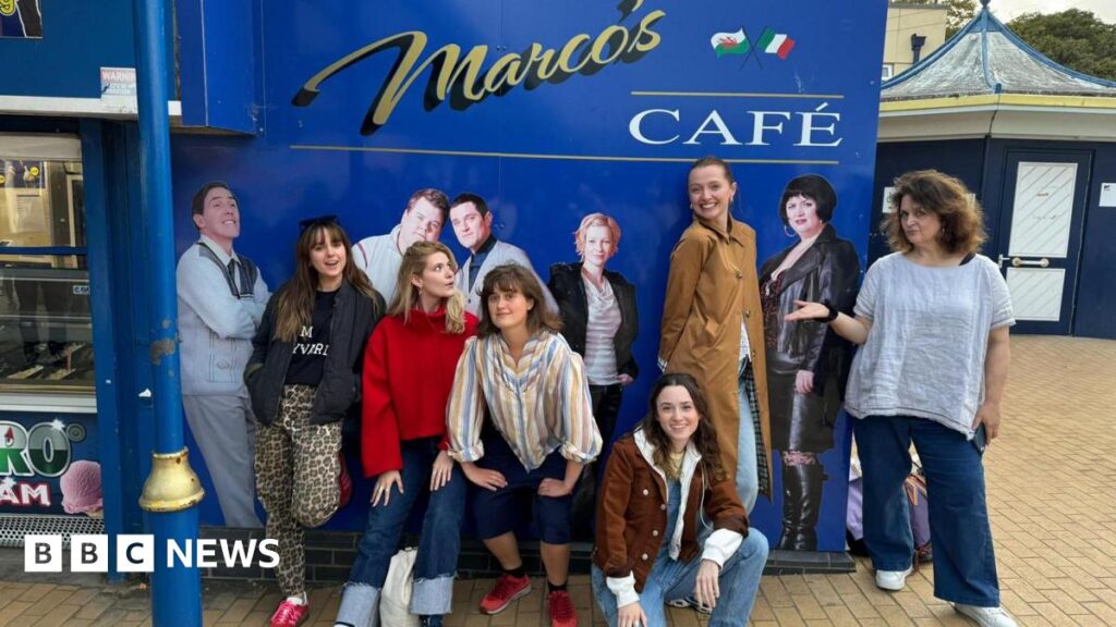 Five women are smiling while standing in front of the blue Marc's Cafe sign at Barry Island, with one woman kneeling on the floor. The sign includes photos of some of the cast of Gavin and Stacey, including Joanna Page as Stacey and Rob Bryden as Uncle Bryn. Ruth Jones is on the right, standing next to a photo of her as Ness.