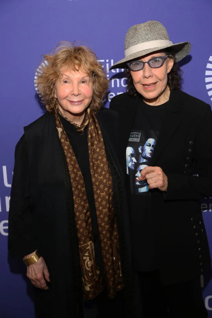 Two smiling individuals pose together. One wears a patterned scarf and gold bracelet; the other is in a hat and glasses with a graphic shirt
