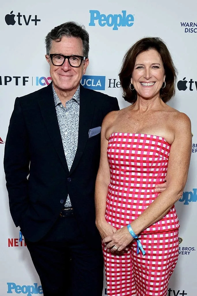 Two people smiling on a red carpet. One wears a suit with a patterned shirt, the other wears a strapless checkered dress