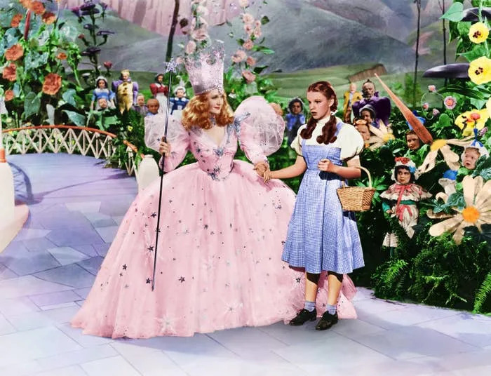Two characters from a classic film: one in a sparkly gown with a crown and wand, the other in a gingham dress holding a basket, surrounded by vibrant scenery