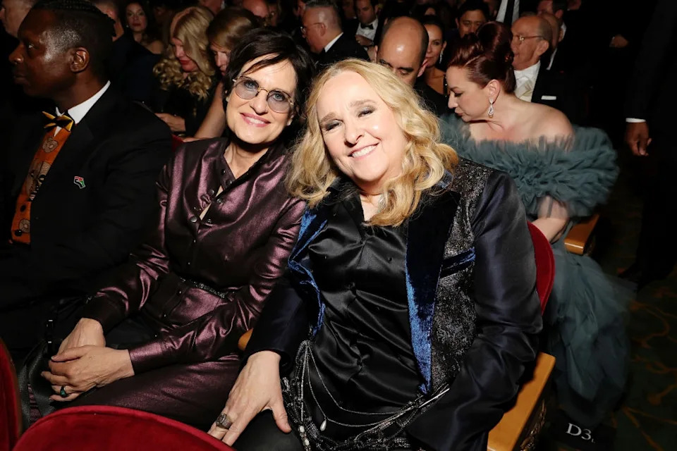 Two people in elegant attire seated closely at an event, surrounded by others. The setting suggests a formal gathering