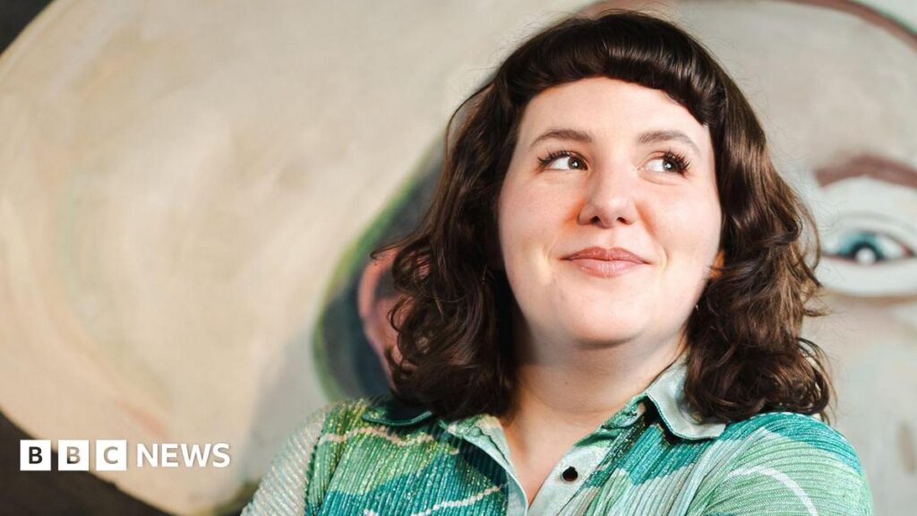 Amanda Dwyer standing in front of a large abstract painting with soft, rounded shapes and muted colours, wearing a patterned green shir