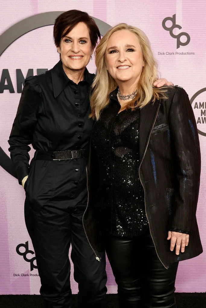 Two individuals pose on the red carpet wearing stylish black outfits; one in a jumpsuit, the other in a sequin top and jacket, smiling at the event
