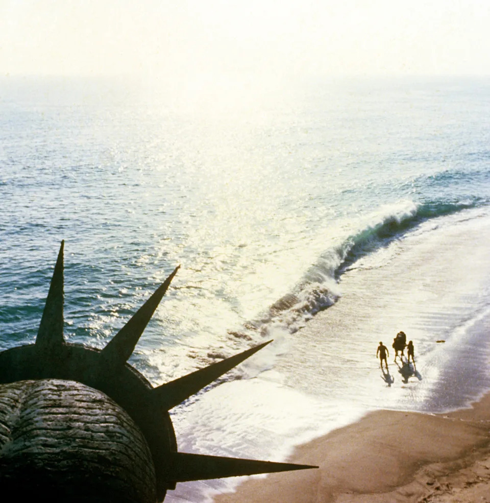 The Statue of Liberty's remains lie on a beach, with people walking in the distance by the surf