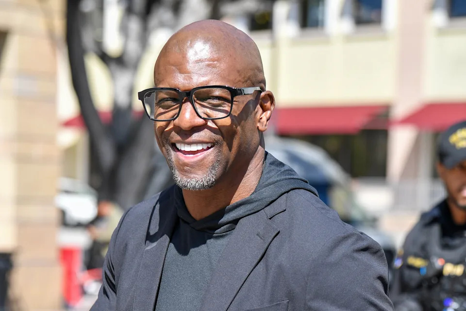 Terry Crews wearing glasses and a black jacket, with people and buildings in the background