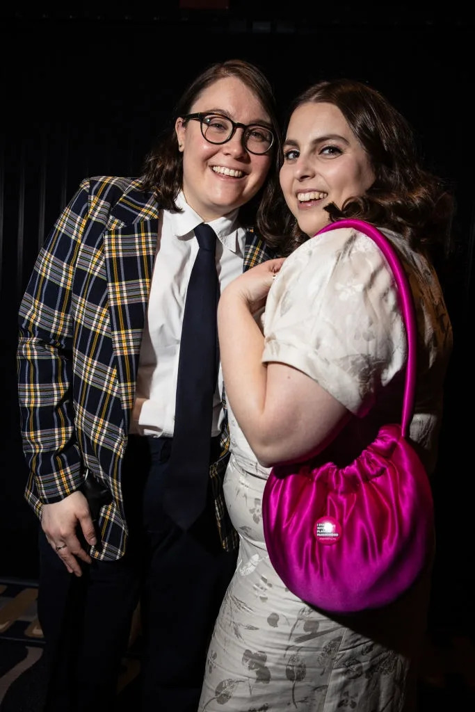 Two people pose together; one in a plaid blazer and tie, the other in a light dress with a bright purse. They smile warmly at the camera