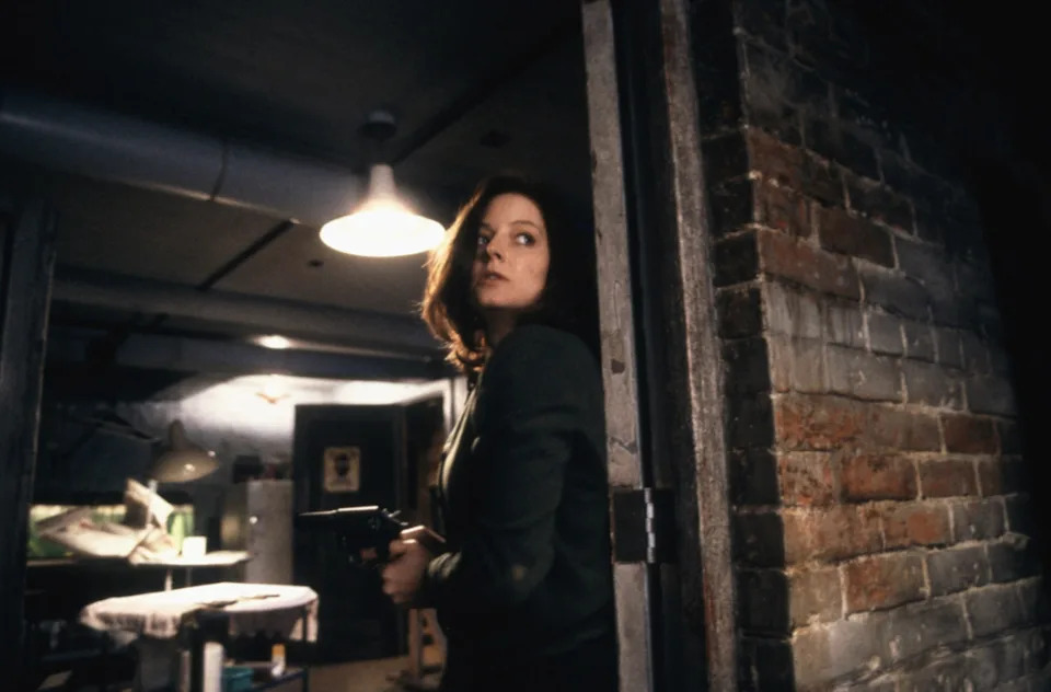 A tense scene with a woman holding a gun, looking alert in a dimly lit, brick-walled basement