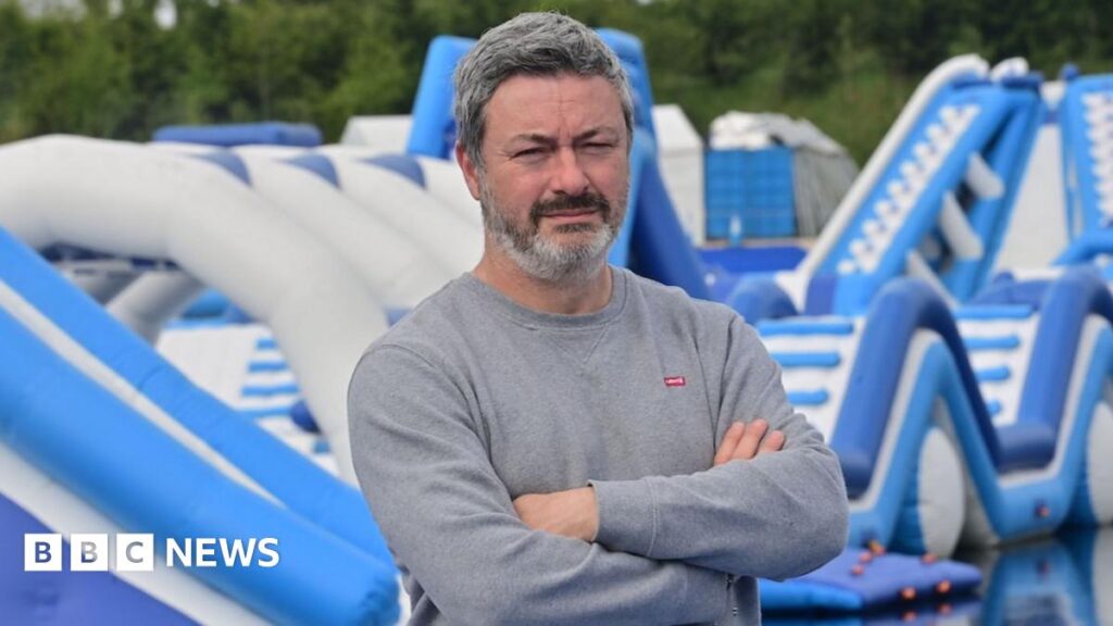 A man in a grey sweatshirt has his arms folded and is standing in front of blue and white water inflatables. He has greying short hair and black and grey beard.