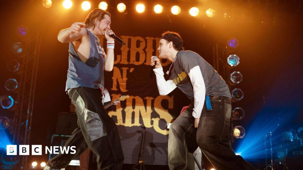 Scheming friends from a council estate – ‘everything’ James McAvoy wanted Two rappers in late 90s/early noughties clothing performing on stage with a backdrop of bright lights and a large sign that reads "Silibil N' Brains"