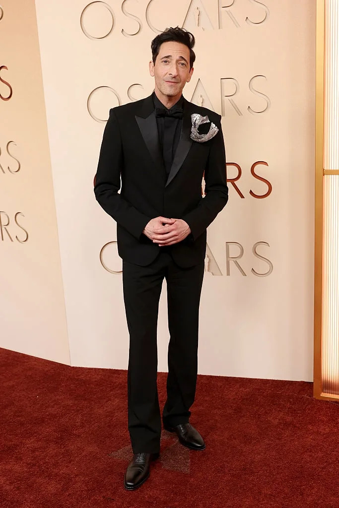 Person in a classic black tuxedo with a unique lapel adornment, posing on a red carpet at a formal event